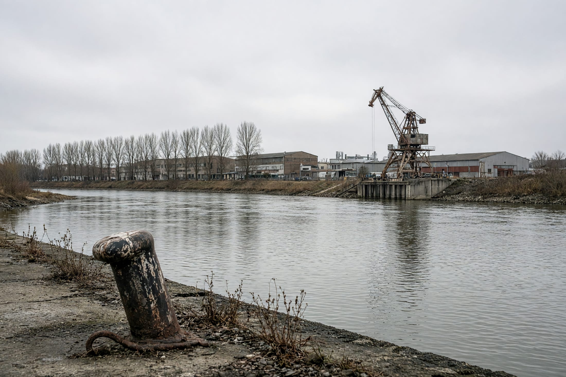 Der Mainbogen bei Fechenheim an einem Novembernachmittag, flaches Licht auf dem Wasser, am Ufer Industriegebäude und ein umgestürzter Poller.